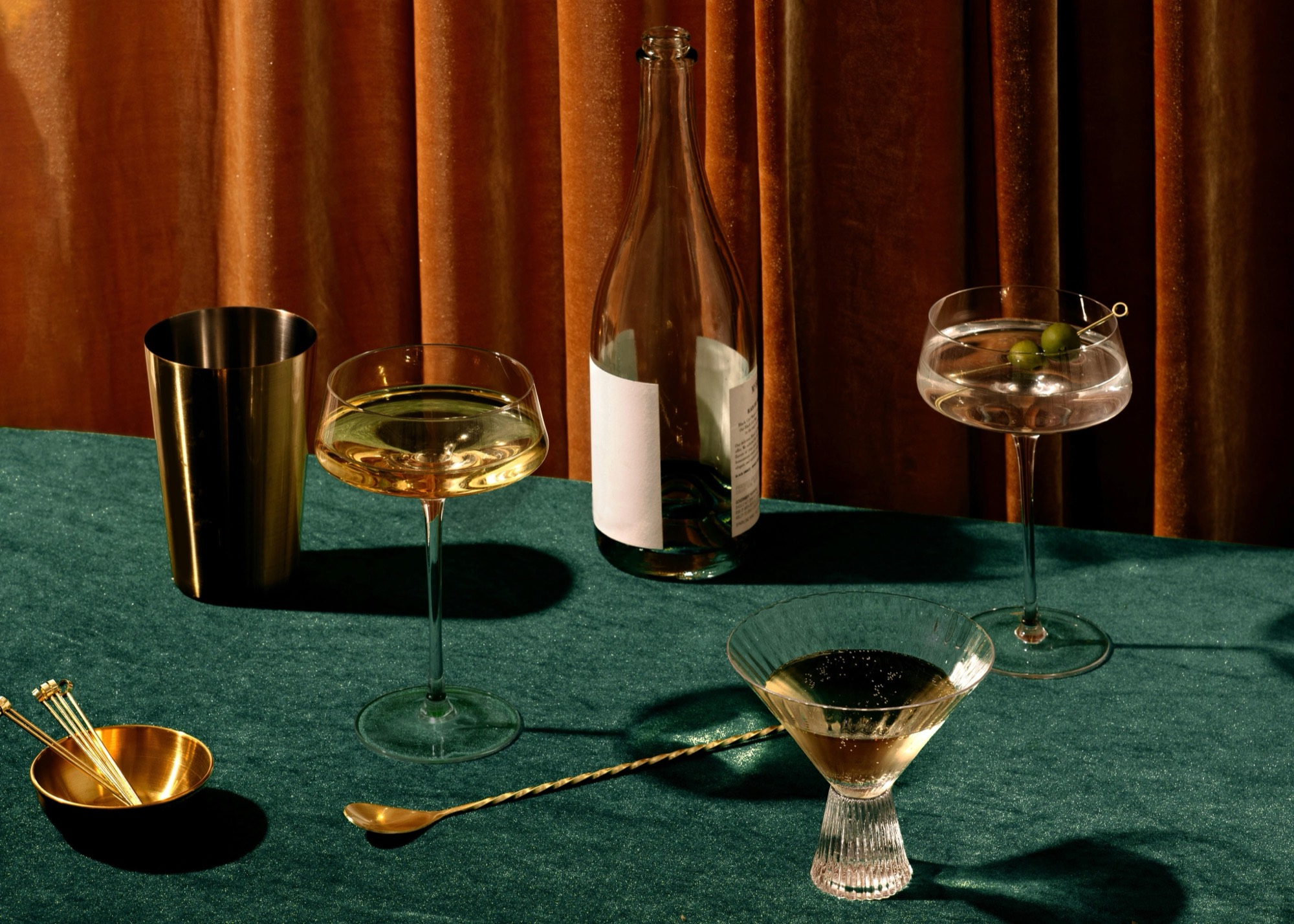 Vesper, martini coupes and a bottle on green velvet against amber curtains